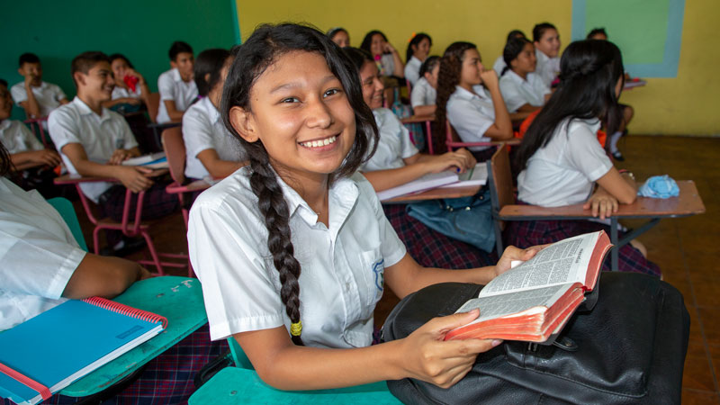 Girl sitting at classroom desk holding open Bible.